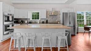 A modern kitchen with white cabinetry, stainless steel appliances, a large island with bar stools, and a view of the outdoors through large windows. - Cape Cod: Gimlet Getaway (Eastham)