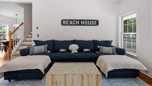 A living room with a dark blue sofa, two matching armchairs, a wooden coffee table, and a 'BEACH HOUSE' sign on the wall. - Cape Cod: Gimlet Getaway (Eastham)