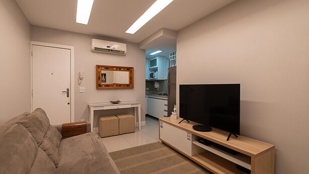 Ein modernes Wohnzimmer mit einer Couch, einem Fernseher auf einem Holzboden und einer Küche im Hintergrund. Eine Klimaanlage ist an der Wand angebracht.