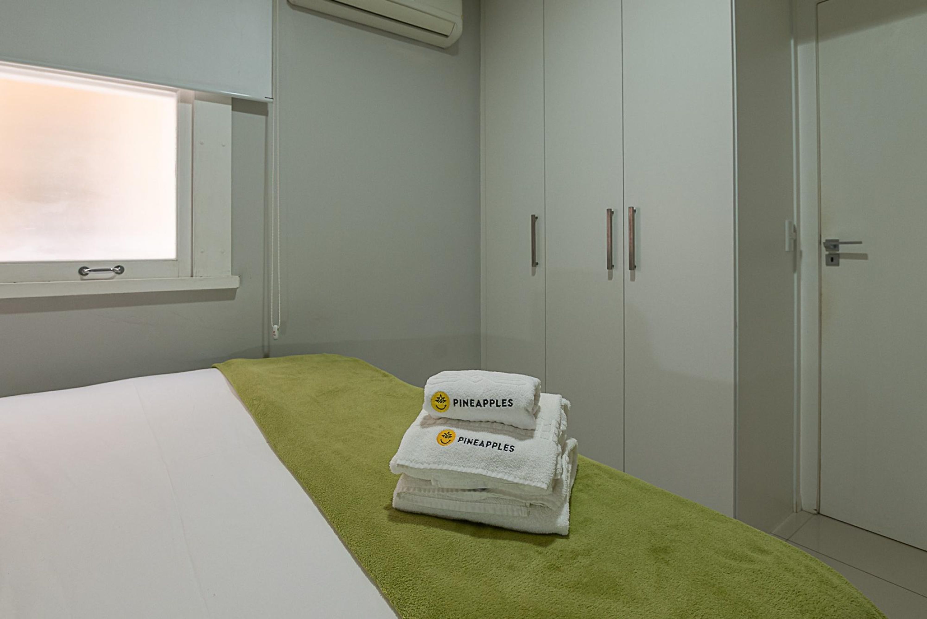 A hotel room with a bed, a green towel, and a stack of white towels with 'Pineapples' printed on them.
