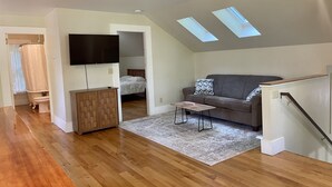 Ein modernes Wohnzimmer mit Holzboden, einem grauen Sofa, einem kleinen Tisch und einem an der Wand befestigten Fernseher. Über dem Sofa befinden sich Dachfenster und rechts eine Treppe.