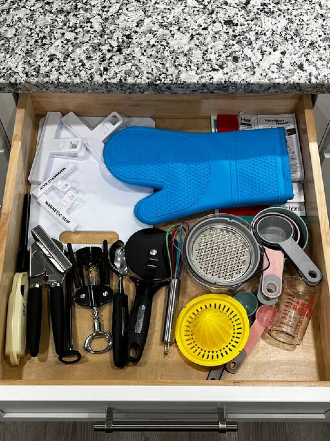 A drawer with kitchen utensils, a blue oven mitt, a yellow colander, and measuring cups.