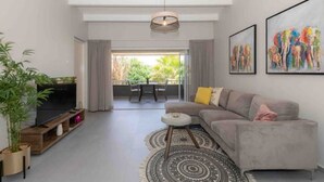 A modern living room with a grey sofa, a TV on a wooden stand, a patterned rug, and a dining area with a table and chairs. - CASA ARTE- Modern Apt+ Car option & Pool (Willemstad)
