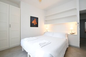 A hotel room with a bed, white walls, a framed picture, two bedside lamps, and a wardrobe.
