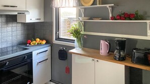 Et kompakt køkken med komfur, ovn og hvide skabe. Der er et vindue med udsigt, en hylde med køkkenudstyr og en vase med blomster på bordpladen.