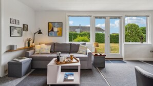 A modern living room with a grey sofa, a coffee table, and a bookshelf. There are large windows with a view of greenery and a house.