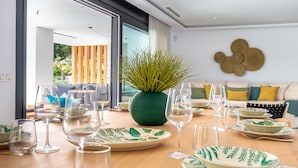 A dining area with a wooden table set for a meal, a green vase with plants, and a sofa with cushions in the background. - All About Playas Andaluzas Villa (Marbella)