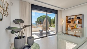 A modern living room with a large sliding glass door leading to a balcony with a view of the sea. There are potted plants, a glass coffee table, and a bookshelf with decorative items. - All About Playas Andaluzas Villa (Marbella)