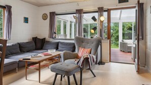 Un salon chaleureux avec un canapé gris, un fauteuil assorti et une table basse en bois. Une grande fenêtre avec des rideaux, une porte donnant sur l’extérieur, et une petite table sur laquelle se trouvent un bol et des livres.