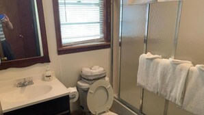 A bathroom with a toilet, sink, and mirror. There is a window with blinds, a shower area, and a towel rack. - Two-Bedroom Apartment (Robbinsville)