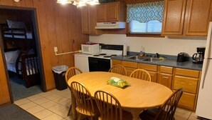 A compact kitchen with wooden cabinets, a microwave, a stove, and a sink. There is a dining table with chairs and a trash bin. - Two-Bedroom Apartment (Robbinsville)