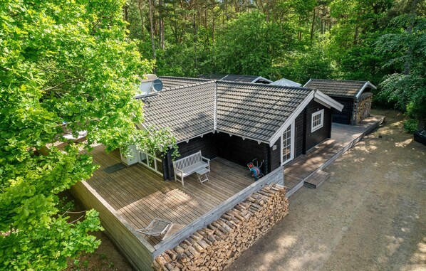 Une cabane avec un toit en pente, une terrasse avec un banc et une pile de bois de chauffage.