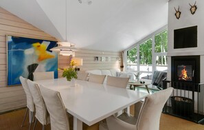 Un espace de salle à manger moderne avec une table et des chaises blanches, un luminaire suspendu et une cheminée où brûle un feu.