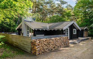 Une cabane avec un toit en tuiles, une terrasse en bois et une pile de bois de chauffage.