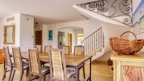Un coin repas avec une table en bois et des chaises en osier, un escalier avec une rampe en fer forgé et un panier tressé posé sur un meuble en bois.