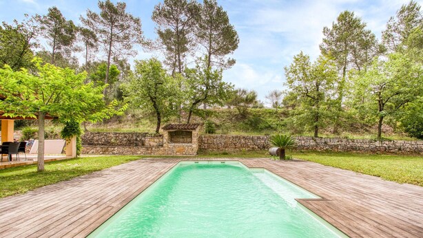 Une piscine rectangulaire entourée d’une terrasse en bois, située dans un jardin aménagé avec des arbres et un mur en pierre.