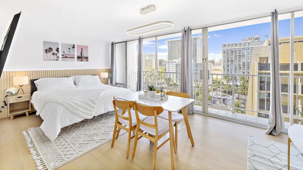 A modern bedroom with a large bed, a dining table, and a city view through floor-to-ceiling windows.