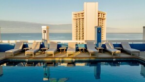 A rooftop pool with lounge chairs and a view of the ocean and a tall building in the background.