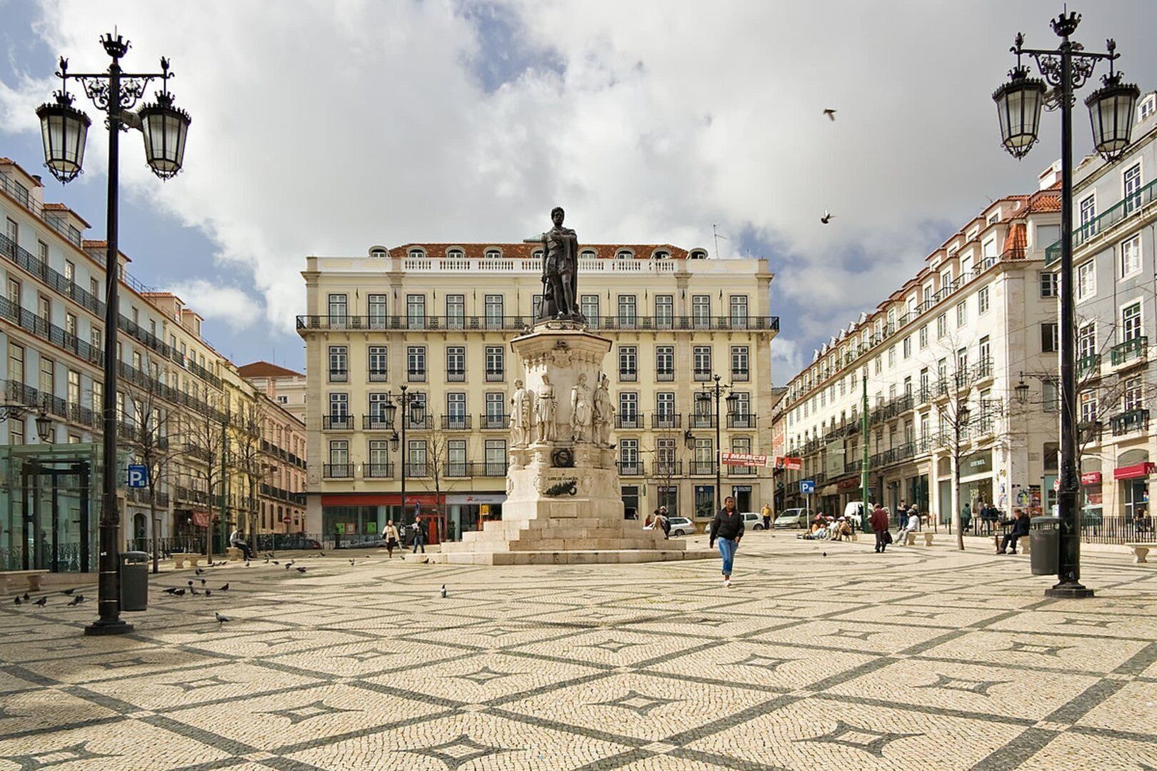 Uma praça pública com um monumento central, cercada por edifícios com varandas e lojas.