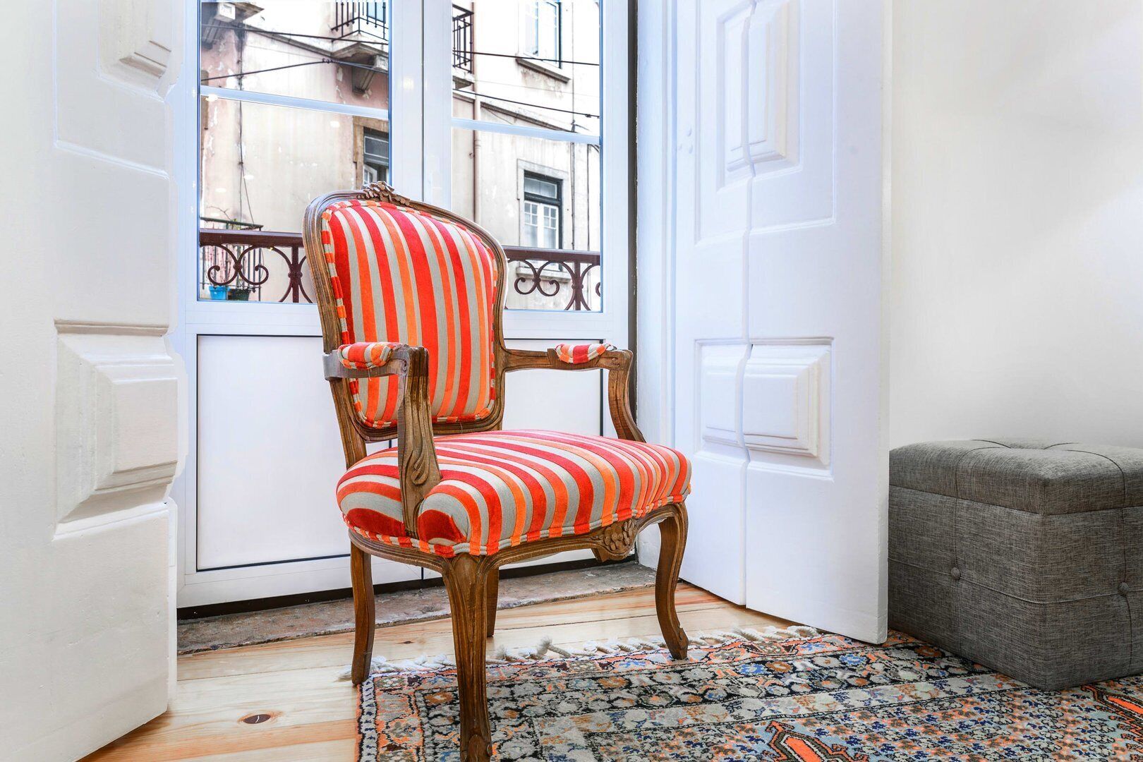 Quarto nos fundos com uma cadeira listrada, um banquinho cinza e um tapete estampado.