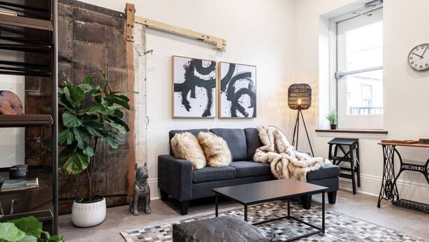 A modern living room with a dark sofa, a coffee table, and a large plant. - Juno Lofts: Memories of Gastown (Stratford)
