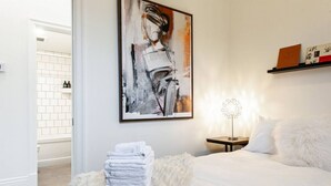 A modern bedroom with a large bed, a white fluffy rug, a painting on the wall, and a bedside table with a lamp. - Juno Lofts: Memories of Gastown (Stratford)
