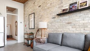 Ein Wohnzimmer mit grauem Sofa, Holzboden, Backsteinwand und Bücherregal mit Büchern und Lampe.