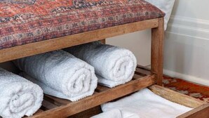 Eine Holzbank mit integriertem Ablagefach, das gerollte Handtücher enthält, ein gemustertes Teppich und ein Bett mit weißer Bettwäsche.