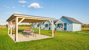 Ein überdachter Außenbereich mit Tisch und Stühlen, ein kleines Gebäude mit Veranda und eine Wiese.