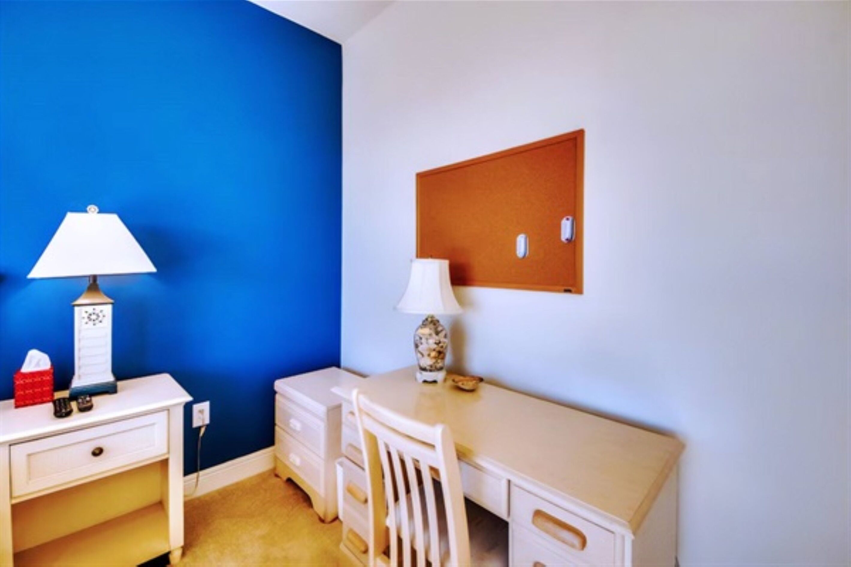 A small room with a blue wall, a white desk with a lamp, a chair, and a corkboard on the wall.