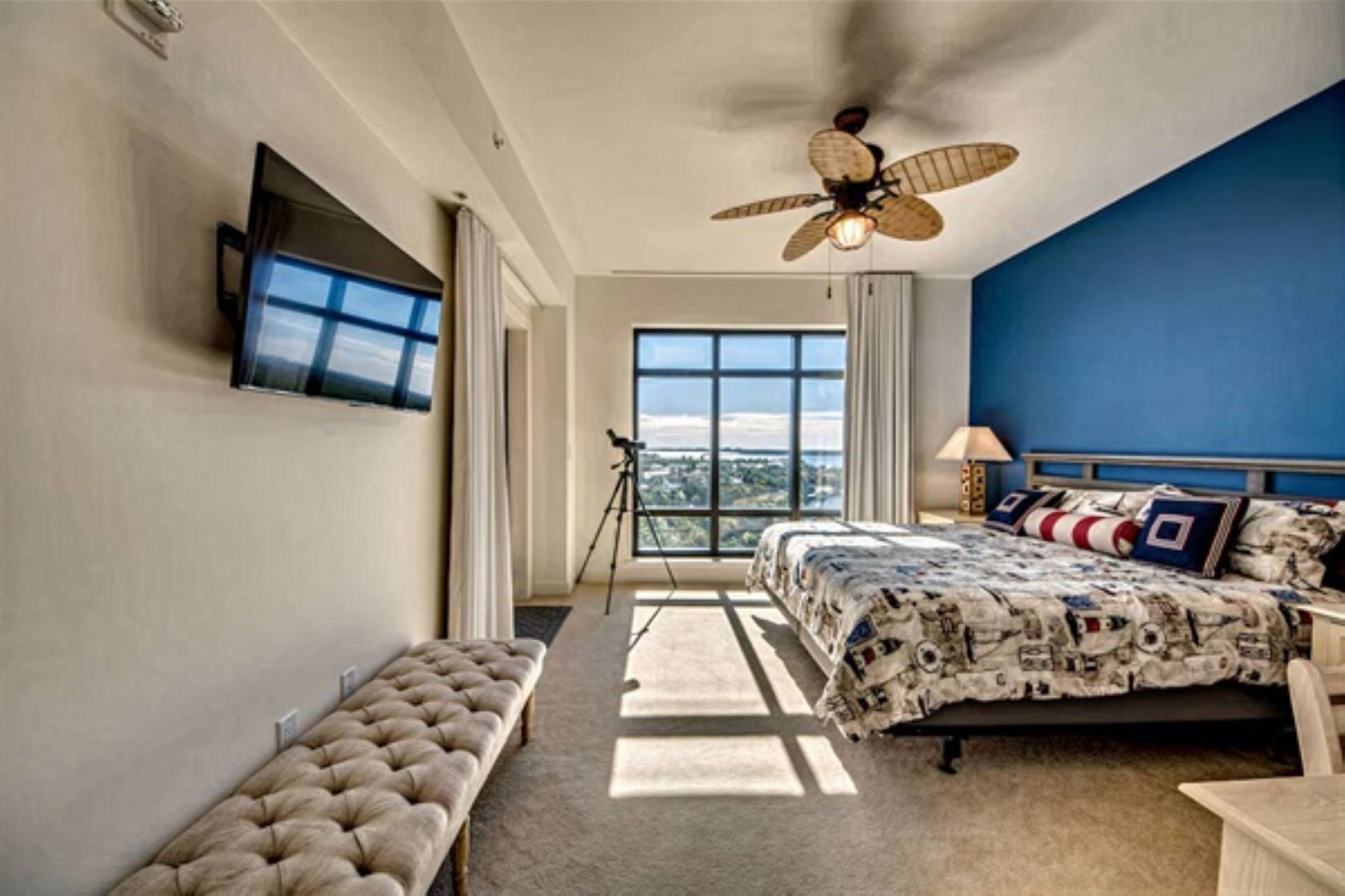 A bedroom with a bed, a bench, a ceiling fan, a window, and a tripod.