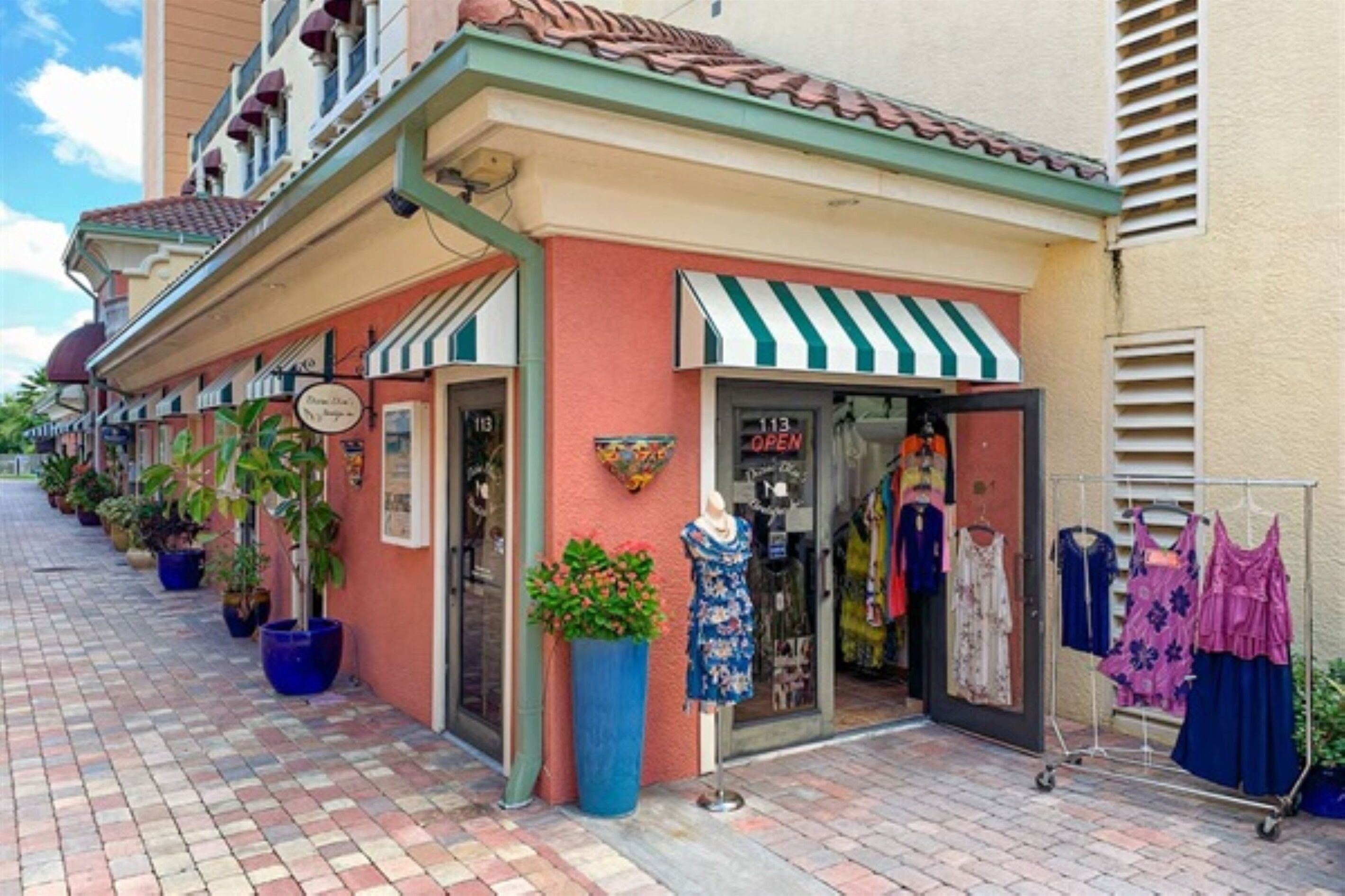 A row of boutique shops with a tiled walkway, potted plants, and a mannequin displaying dresses.