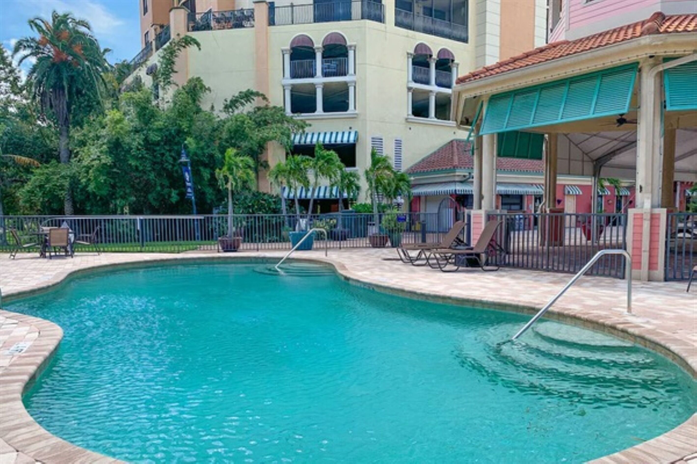 A swimming pool with a tiled deck, surrounded by a fence, and adjacent to a building with a covered patio area.