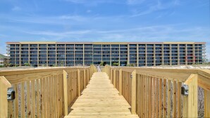 A wooden boardwalk leading to a multi-story hotel with a flat roof and numerous windows. - UPDATED Beach Front Condo - Holiday Isle 302 - Pool, Sauna, Hot Tub, Grill, W/D (Dauphin Island)
