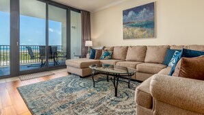 A living room with a glass coffee table, beige sofa, and a view of the ocean through large sliding glass doors. - UPDATED Beach Front Condo - Holiday Isle 302 - Pool, Sauna, Hot Tub, Grill, W/D (Dauphin Island)