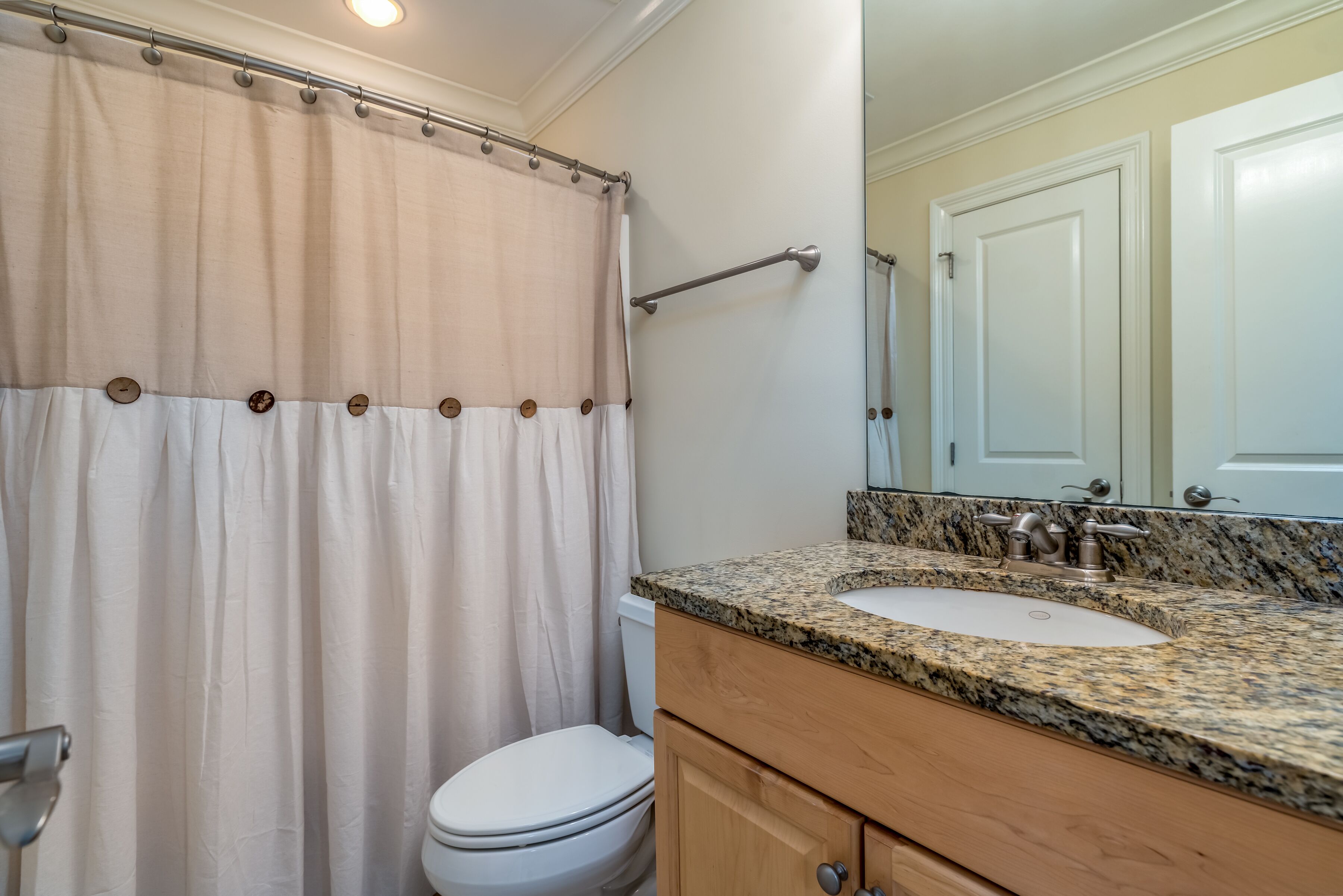 A bathroom with a beige shower curtain, a white toilet, a granite countertop sink, and wooden cabinets.