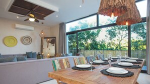 A dining area with a wooden table set for six, large windows, and a ceiling fan. - Manor Tulum by The Spot Rentals (Tulum)