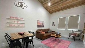 A cozy living room with a wooden ceiling, a brown sofa, a dining table with chairs, and a pink rug.