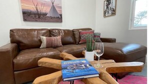 A living room with a brown leather sofa, a wooden coffee table with a book and a glass, a cactus plant, and a painting of a desert scene.