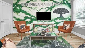 Een woonkamer met een muurschildering getiteld 'THE MELANATED WAY', twee bruine leren fauteuils, een salontafel en een aan de muur bevestigde televisie.