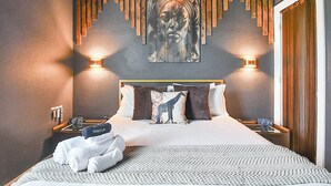 Een moderne hotelkamer met een groot bed, een aan de muur bevestigd kunstwerk, twee nachtkastjes met klokken, en een opgemaakt bed met handdoeken en een kamerkleed met een patroon.