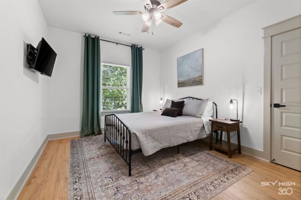 A bedroom with a bed, a ceiling fan, a TV mounted on the wall, a window with curtains, and a door.