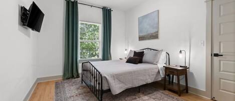 A bedroom with a bed, a ceiling fan, a TV mounted on the wall, a window with curtains, and a door.