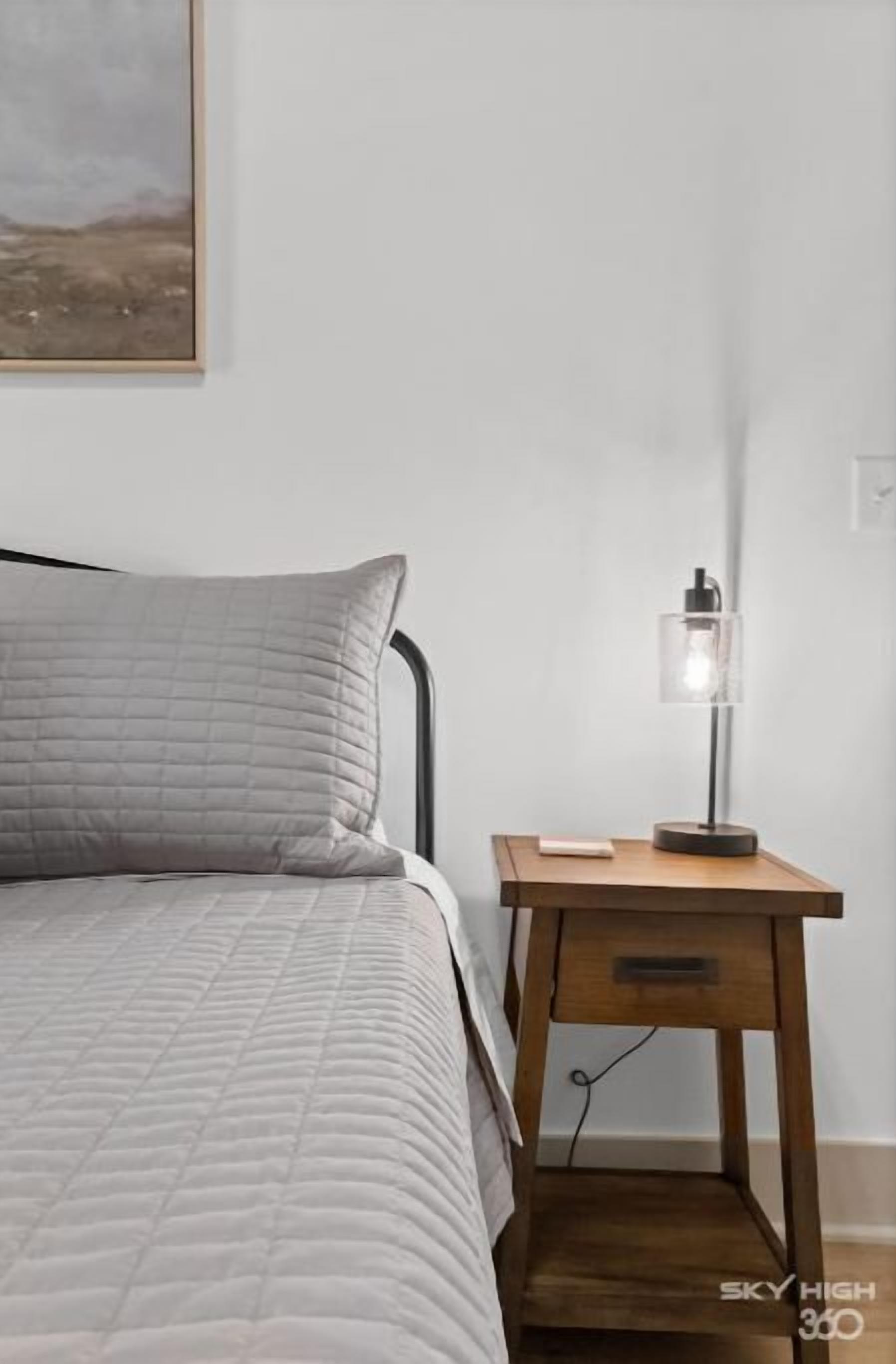 En seng med et quiltet gråt dynedække og et natbord i træ med en lampe.