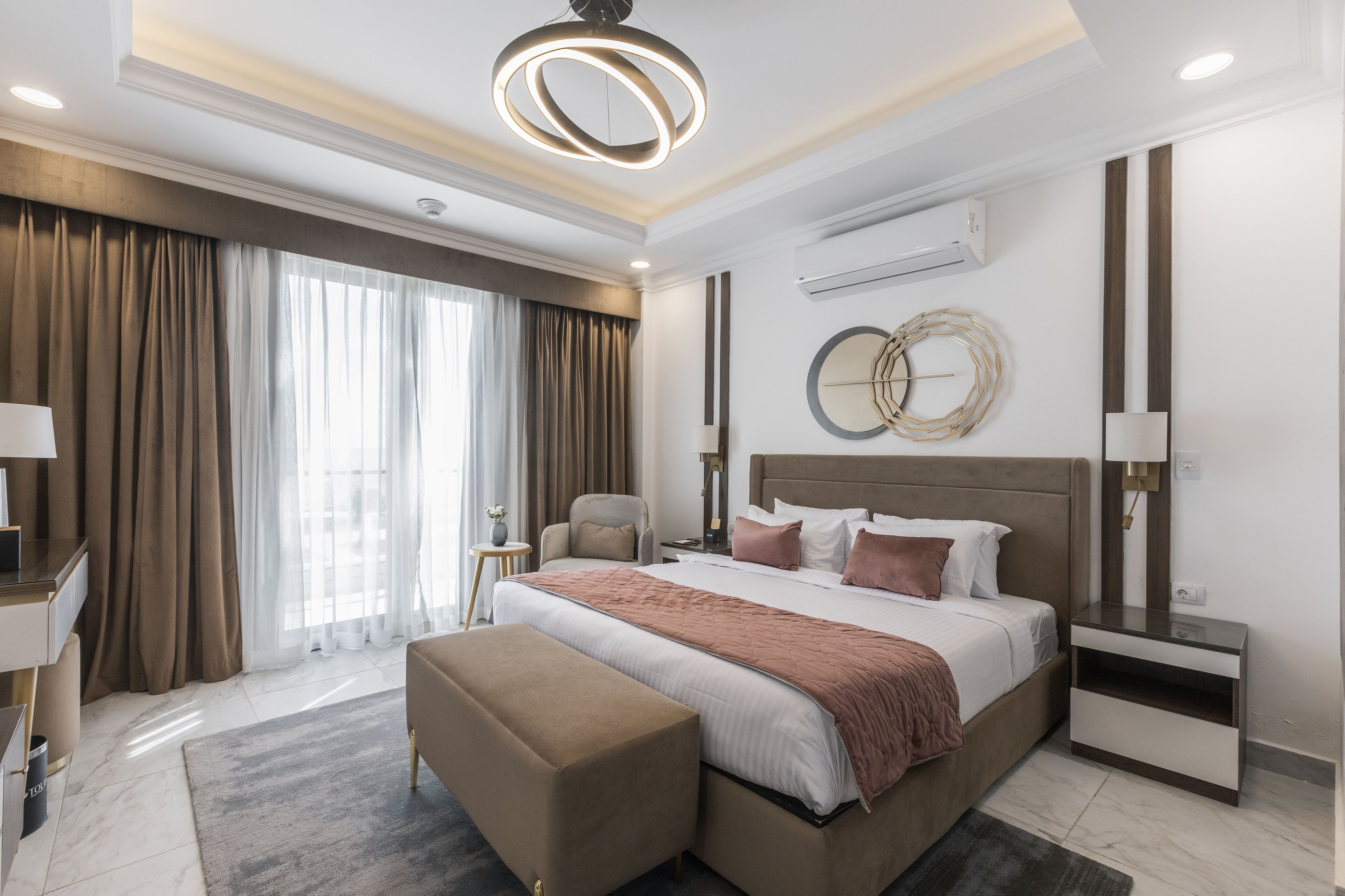 A modern hotel room with a large bed, a bedside table, a chair, a desk, and a large window with curtains.