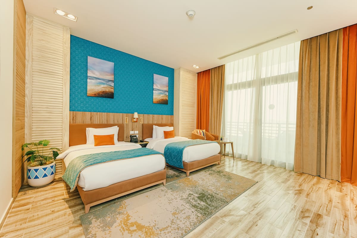 A hotel room with two beds, a large window with curtains, a wall-mounted artwork, and a wooden floor.