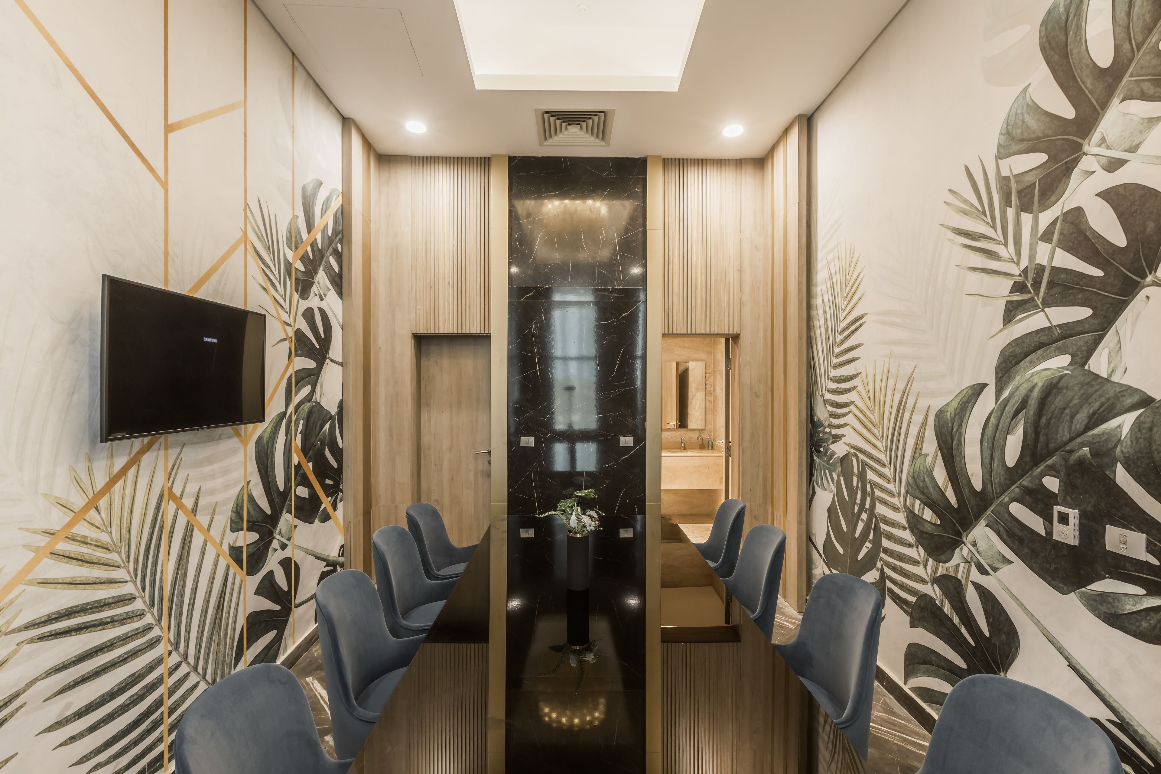 A conference room with a long table, blue chairs, a TV on the wall, and tropical leaf wall art.