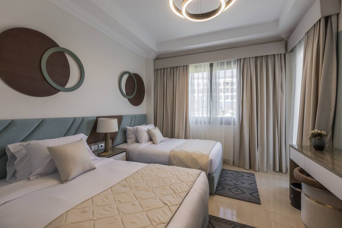 A hotel room with two beds, a desk, a chair, a lamp, and a large window with curtains.