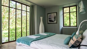 Un dormitorio con un ventanal grande, un florero blanco, una cama con una manta azul turquesa y una almohada con estampado geométrico.