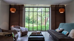 Una sala de estar con sofá, sillón y mesa de centro, una ventana grande con vistas a zonas verdes, y dos lámparas colgantes.
